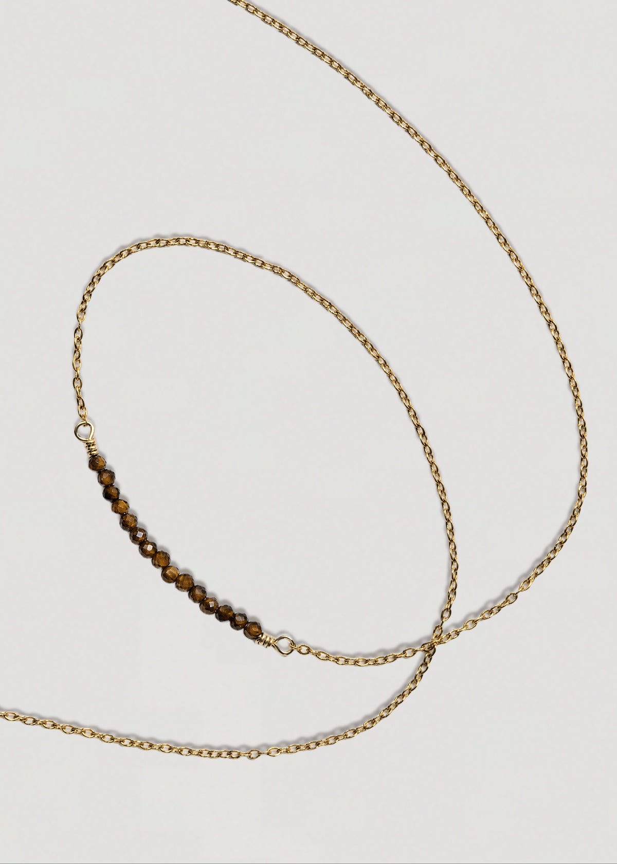 A delicate gold chain necklace featuring a small central bar of brown tiger's eye beads, displayed on a light gray background.