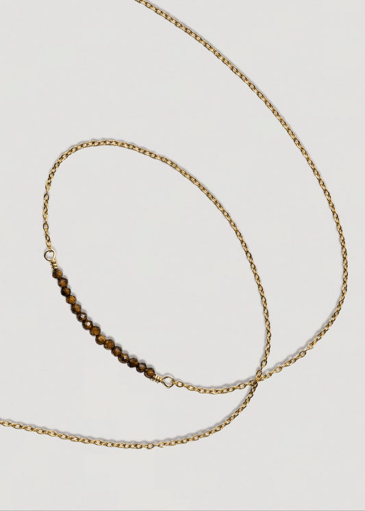A delicate gold chain necklace featuring a small central bar of brown tiger's eye beads, displayed on a light gray background.